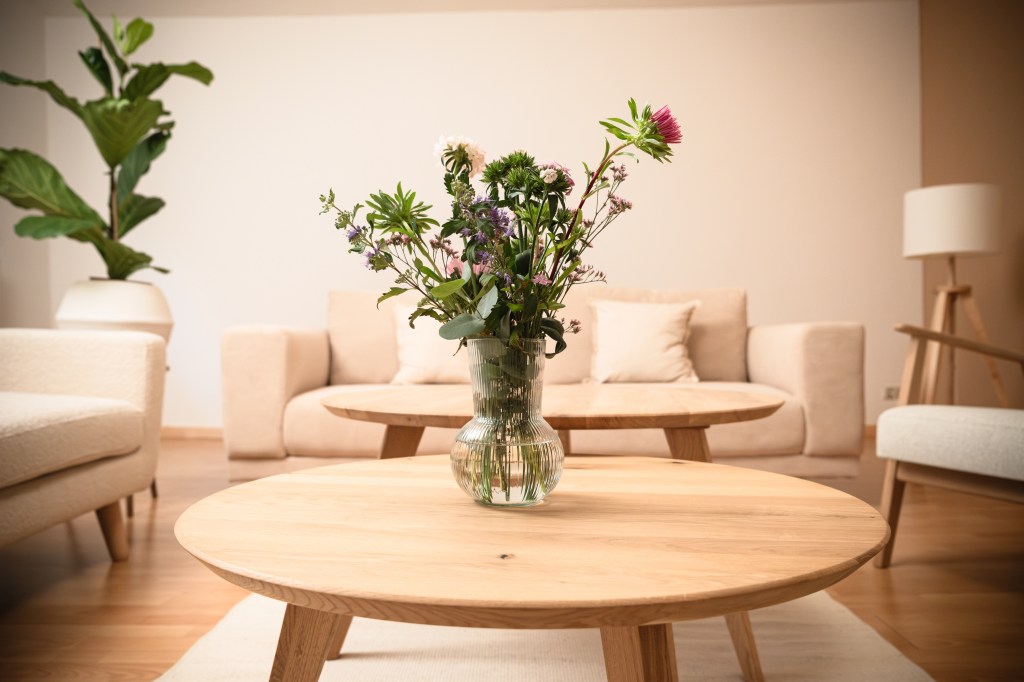 Blumenstrauß in Glasvase auf rundem Holztisch im Gruppentherapieraum, im Hintergrund ein beiges Sofa, helle Sessel und eine große Zimmerpflanze
