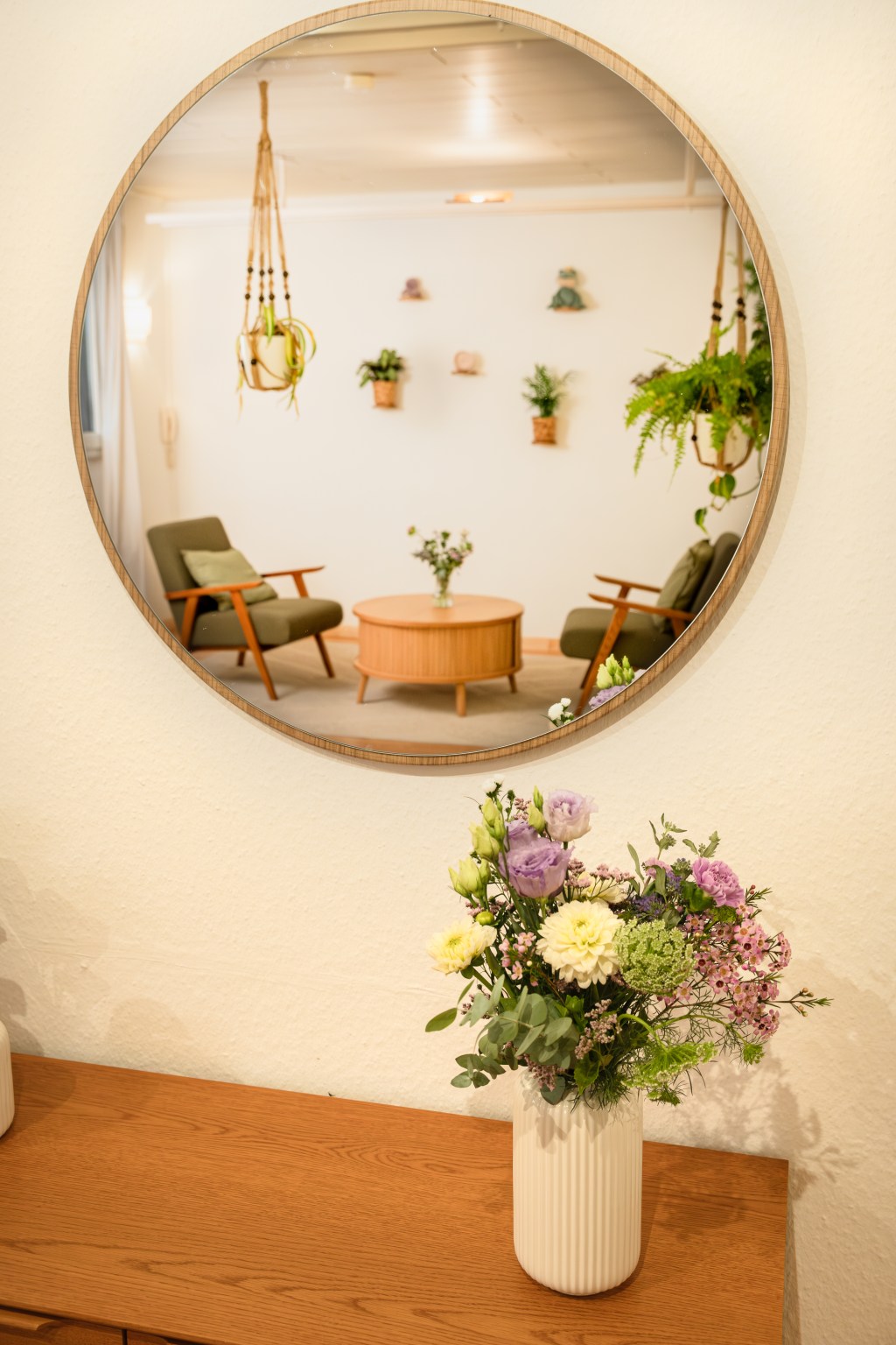 Blumenstrauß in Vase auf Holzkommode, darüber ein runder Spiegel, in dem ein Einzeltherapieraum mit zwei grünen Sesseln, rundem Holztisch und Wandpflanzen zu sehen ist