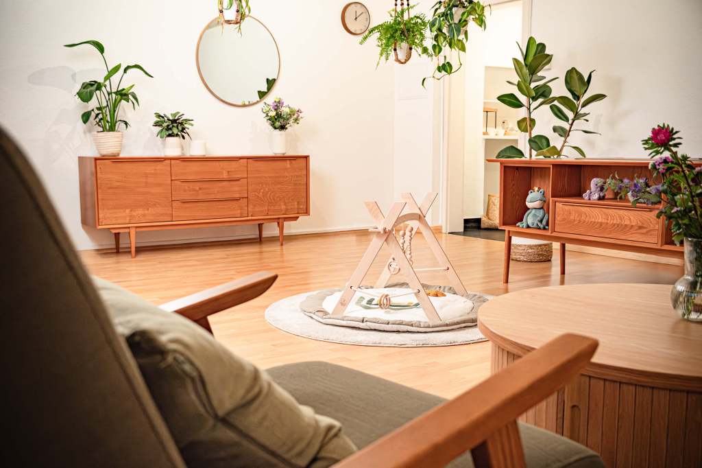 Einzeltherapieraum mit Holzspielbogen auf rundem Teppich, links und rechts Holzkommoden mit Pflanzen und Deko, im Hintergrund eine offene Tür, im Vordergrund ein Sessel und ein Holztisch