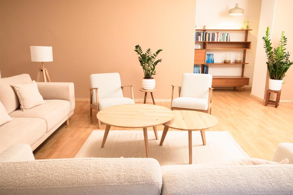 Gemütlich eingerichteter Gruppentherapieraum mit cremefarbenem Sofa, zwei hellen Sesseln, runden Holztischen und mehreren Zimmerpflanzen; im Hintergrund ein Regal mit Büchern und Aktenordnern